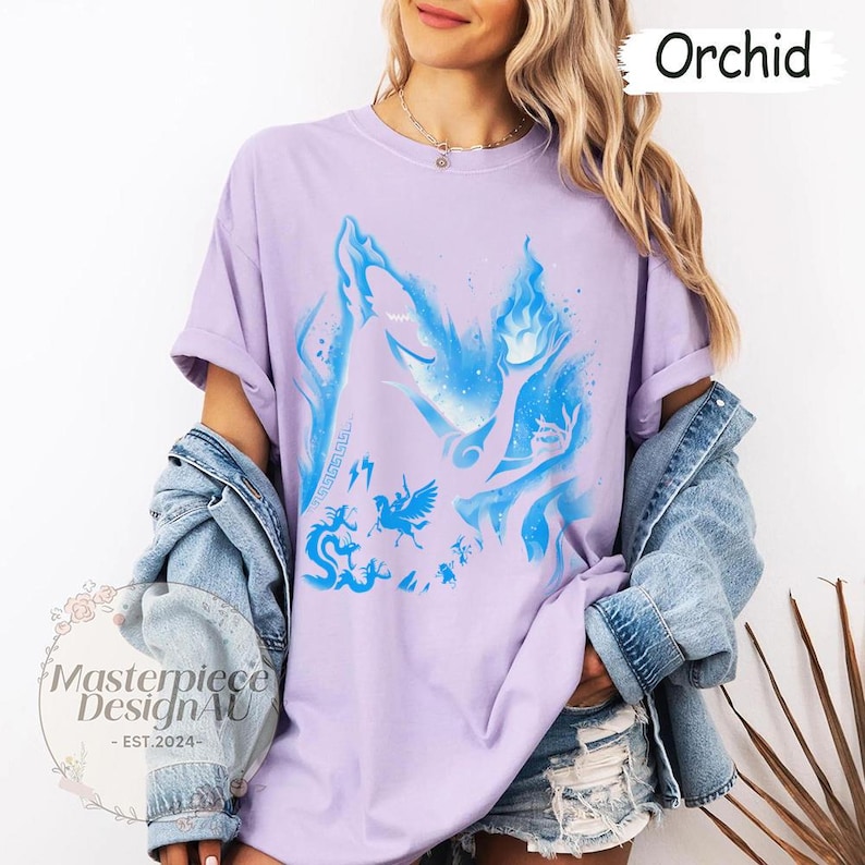 May include: Lilac t-shirt with a blue illustration of mythical figures and flames. The shirt has rolled-up sleeves and is worn with a denim jacket. The word "Orchid" is displayed in the top right corner.