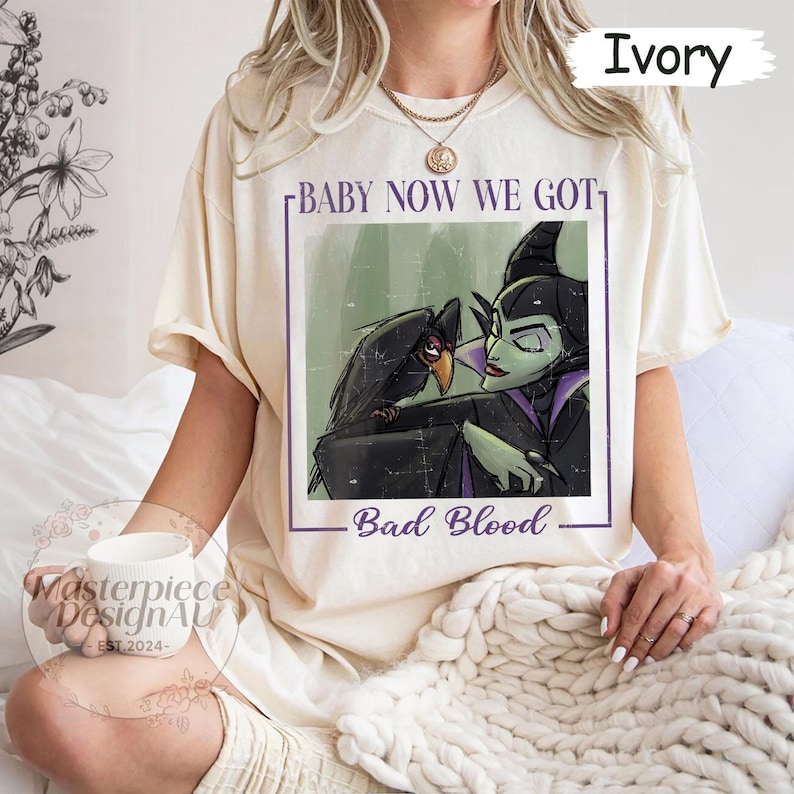 May include: Ivory-colored t-shirt featuring a graphic of a raven and a green-skinned character with the text "BABY NOW WE GOT" and "Bad Blood". The shirt is a relaxed fit.