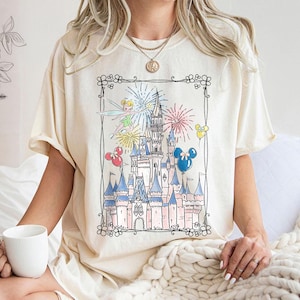 May include: Cream-colored t-shirt featuring a whimsical illustration of a fairytale castle with fireworks, Tinkerbell, and Mickey Mouse balloons. The castle is detailed with pink and blue accents, set within a decorative black border. A casual, comfortable tee.