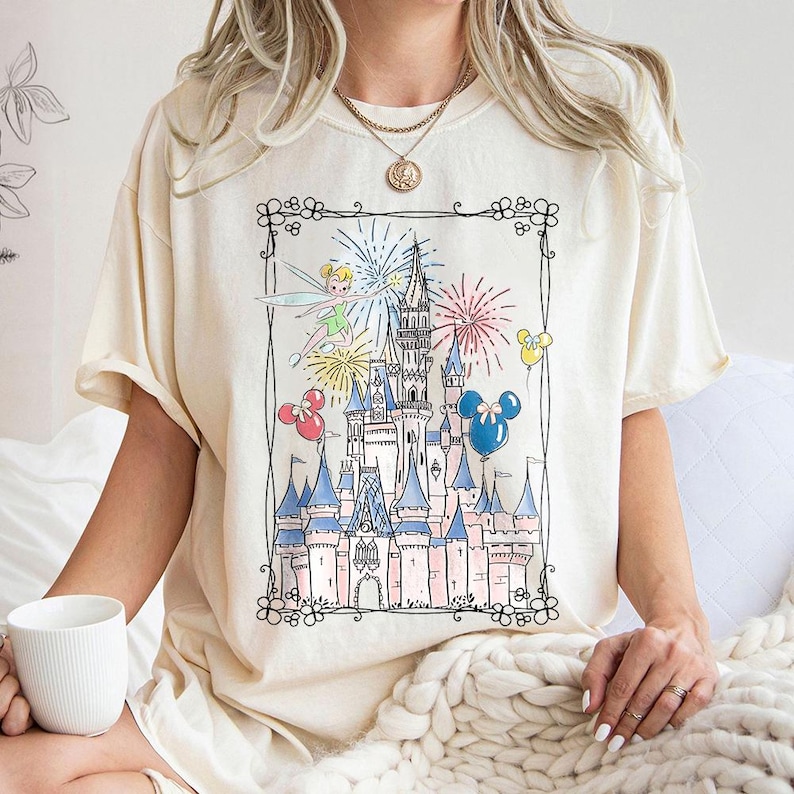 May include: Cream-colored t-shirt featuring a whimsical illustration of a fairytale castle with fireworks, Tinkerbell, and Mickey Mouse balloons. The castle is detailed with pink and blue accents, set within a decorative black border. A casual, comfortable tee.