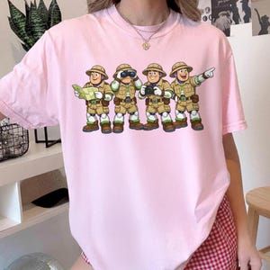 Peut inclure: T-shirt rose clair avec une illustration de dessin anim&eacute; de quatre explorateurs. Les explorateurs portent des tenues kaki, des chapeaux et des bottes. L'un tient une carte, un autre des jumelles, un troisi&egrave;me un appareil photo et le dernier pointe vers l'avant.