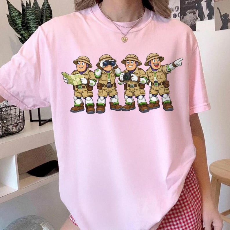 Peut inclure: T-shirt rose clair avec une illustration de dessin anim&eacute; de quatre explorateurs. Les explorateurs portent des tenues kaki, des chapeaux et des bottes. L'un tient une carte, un autre des jumelles, un troisi&egrave;me un appareil photo et le dernier pointe vers l'avant.