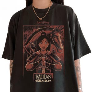 May include: Black t-shirt featuring a graphic of Mulan holding a sword, with a horse and dragon in the background. The text "Walt Disney Pictures Presents" and "Mulan" are also visible. The shirt has a vintage look.