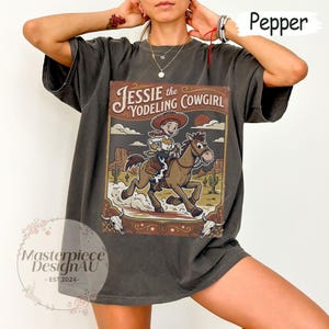 May include: A charcoal gray t-shirt featuring a graphic print of a cowgirl riding a horse in a desert landscape. The text reads Jessie the Yodeling Cowgirl. The design includes cacti, a sun, and two cow skulls at the bottom corners of the frame.