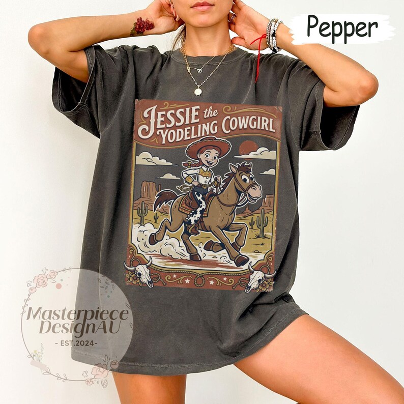 May include: A charcoal gray t-shirt featuring a graphic print of a cowgirl riding a horse in a desert landscape. The text reads Jessie the Yodeling Cowgirl. The design includes cacti, a sun, and two cow skulls at the bottom corners of the frame.