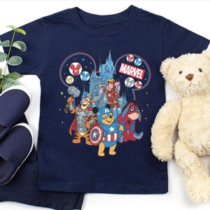 Może przedstawiać: Granatowa koszulka z nadrukiem łączącym Disney i Marvel. Grafika przedstawia postacie z Kubusia Puchatka jako Avengers, z logo Marvela i zamkiem w tle. Widoczny jest pluszowy miś i niebieskie buty.