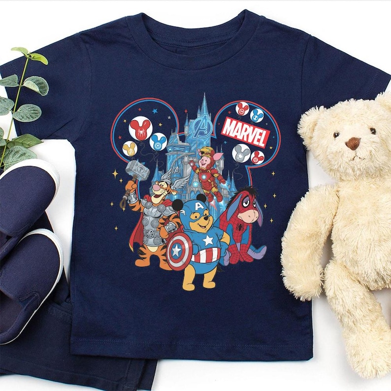 Może przedstawiać: Granatowa koszulka z nadrukiem łączącym Disney i Marvel. Grafika przedstawia postacie z Kubusia Puchatka jako Avengers, z logo Marvela i zamkiem w tle. Widoczny jest pluszowy miś i niebieskie buty.