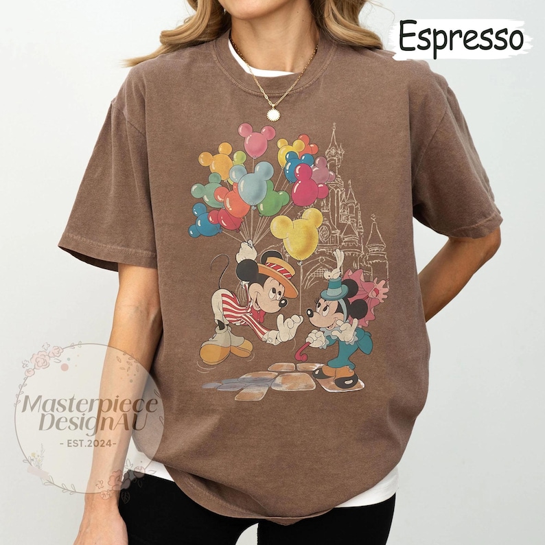 May include: A brown t-shirt featuring a vintage-style graphic of Mickey Mouse and Minnie Mouse holding balloons in front of a castle. The text "Espresso" is in the top right corner of the image.