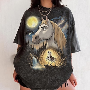 May include: A charcoal grey acid-wash t-shirt featuring a large graphic print of a horse head against a night sky with a full moon and a shooting star, above a smaller horse standing on a rocky cliff near a waterfall and a glowing cave entrance.