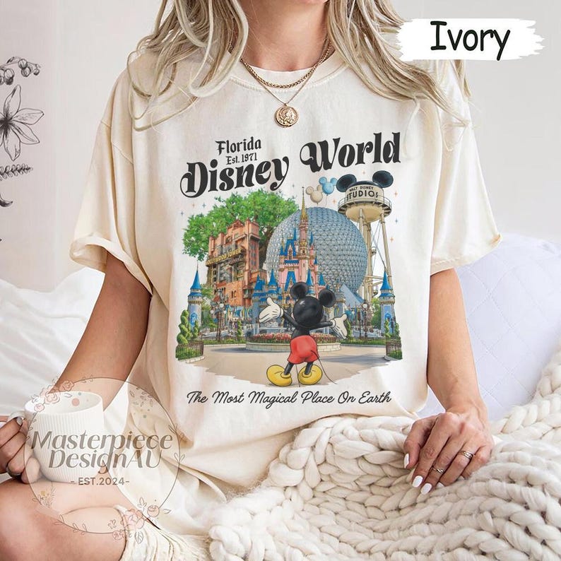 May include: Ivory-colored t-shirt featuring a colorful graphic of Disney World with Mickey Mouse. The text "Florida Disney World" and "The Most Magical Place On Earth" are also displayed. The shirt is a relaxed fit.