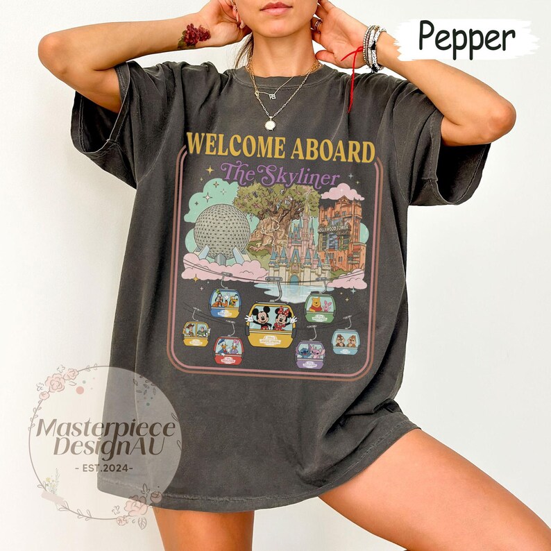 May include: Dark gray t-shirt with a colorful graphic featuring the text "WELCOME ABOARD The Skyliner." The design includes a theme park scene with gondolas, a castle, and other iconic elements. The shirt has a vintage, worn look.