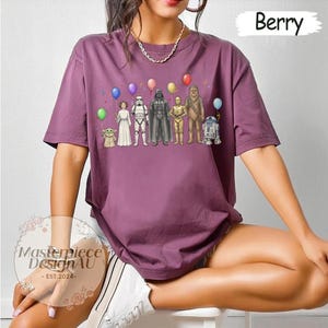 May include: Berry-coloured t-shirt with a Star Wars character graphic and balloons. The graphic includes characters such as Yoda, Princess Leia, Darth Vader, and R2-D2. The shirt has short sleeves and a relaxed fit. The text "Masterpiece DesignAU - EST.2024-" is also visible.