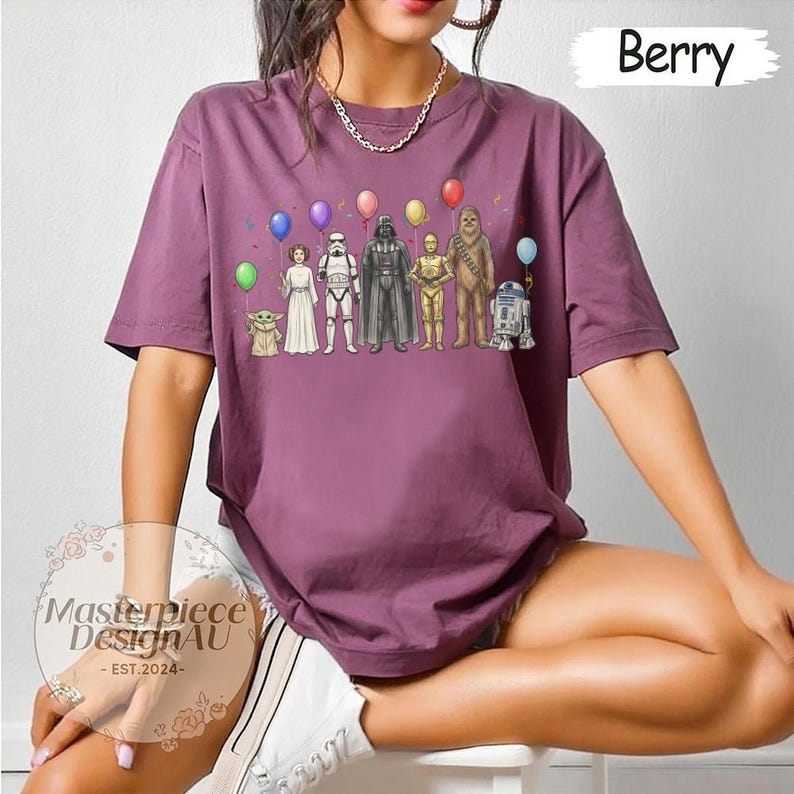 May include: Berry-coloured t-shirt with a Star Wars character graphic and balloons. The graphic includes characters such as Yoda, Princess Leia, Darth Vader, and R2-D2. The shirt has short sleeves and a relaxed fit. The text "Masterpiece DesignAU - EST.2024-" is also visible.