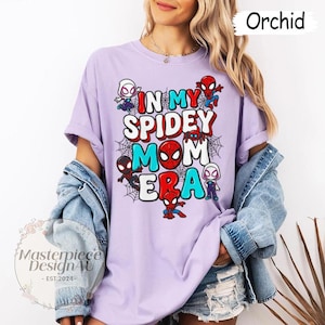 Puede incluir: Camiseta color orqu&iacute;dea con el texto "IN MY SPIDEY MOM ERA" en rojo, blanco y turquesa. Personajes de Spider-Man y telara&ntilde;as rodean el texto. La camiseta es de estilo casual.