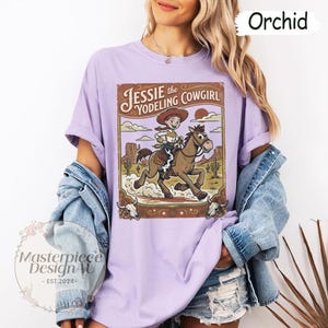 May include: An orchid-colored t-shirt featuring a graphic print of a cowgirl riding a horse in a desert landscape. The text reads Jessie the Yodeling Cowgirl. The design includes cacti, mesas, and two cow skulls at the bottom corners of the frame.