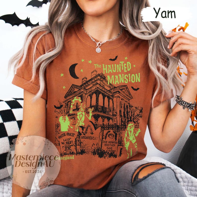 May include: A burnt orange t-shirt featuring a graphic of a haunted mansion with the text "The Haunted Mansion". The design includes bats, a crescent moon, and tombstones. The shirt also has the text "Master Gracey Laid to Rest" and "Dear Departed Brother Dave".