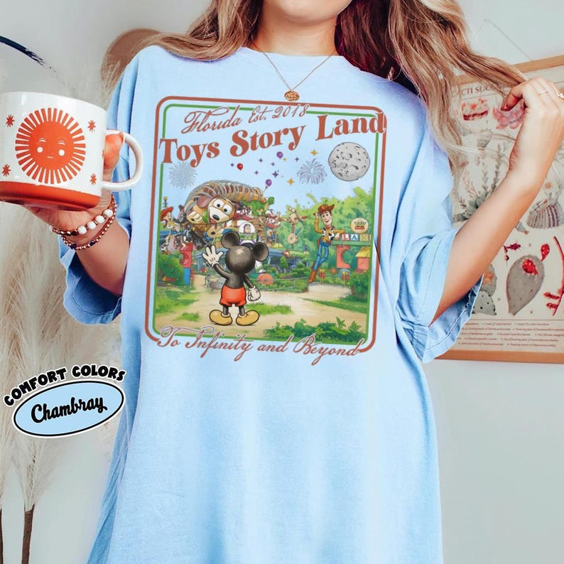 May include: Pale blue t-shirt with a vibrant graphic depicting a theme park scene with Mickey Mouse, Woody, and other characters. The graphic includes the text "Florida Est. 2018 Toys Story Land" and "To Infinity and Beyond". The shirt is a Comfort Colors Chambray.