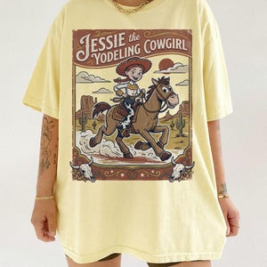 May include: A pale yellow t-shirt featuring a graphic illustration of a cowgirl riding a brown horse in a desert landscape. The text reads Jessie the Yodeling Cowgirl. The design includes cacti, clouds, and two cow skulls in the corners.