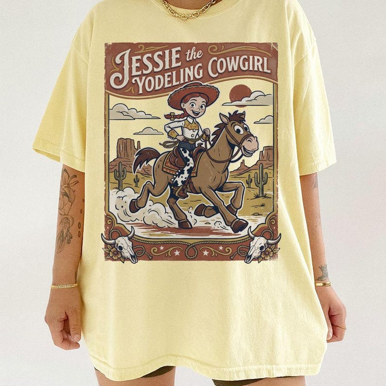 May include: A pale yellow t-shirt featuring a graphic illustration of a cowgirl riding a brown horse in a desert landscape. The text reads Jessie the Yodeling Cowgirl. The design includes cacti, clouds, and two cow skulls in the corners.