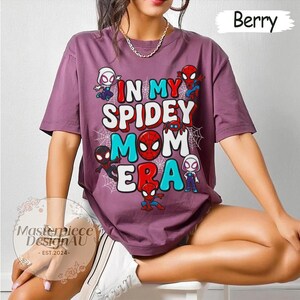 Puede incluir: Camiseta color bayas con el texto "IN MY SPIDEY MOM ERA" y personajes de Spider-Man. La camiseta tiene cuello redondo y mangas cortas. El dise&ntilde;o incluye telara&ntilde;as y varias figuras de Spider-Man en rojo, blanco y azul.