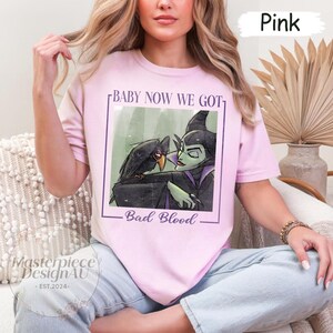 May include: Pink t-shirt featuring a graphic of a raven and a green-skinned character with the text "BABY NOW WE GOT" and "Bad Blood". The shirt has a crew neck and short sleeves. The text "Masterpiece DesignAU - EST.2024-" is also visible.