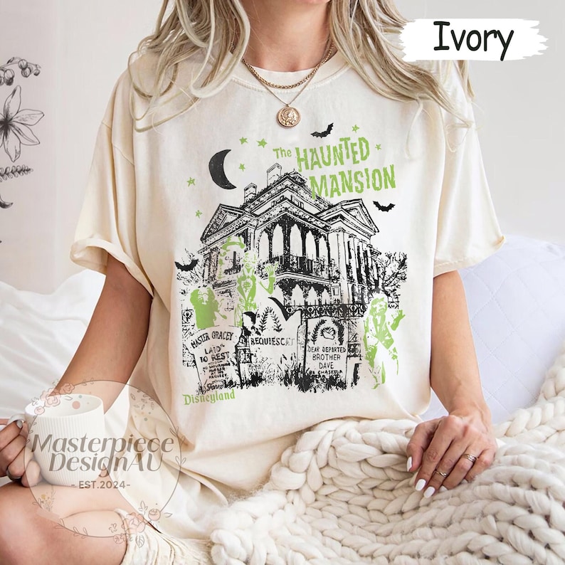 May include: Ivory-colored t-shirt featuring a graphic of The Haunted Mansion, with a spooky house design, moon, stars, and bats. The text "The Haunted Mansion" is above the house, and "Disneyland" is below. The shirt also has the text "Master Gracey Laid to Rest" and "Dear Departed Brother Dave".