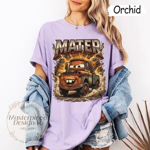 May include: Orchid-colored t-shirt featuring a graphic of Mater from the Cars movie. The design includes the word "MATER" in a metallic font, with a cartoon image of Mater. The shirt is part of a clothing collection.