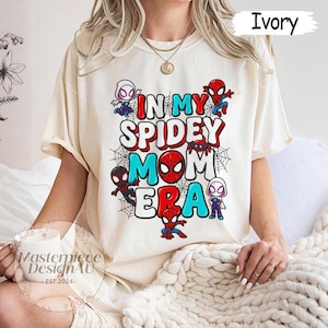 Puede incluir: Camiseta color marfil con el texto "IN MY SPIDEY MOM ERA", personajes de Spider-Man y telara&ntilde;as. El dise&ntilde;o utiliza acentos rojos, azules y blancos. La camiseta es de estilo casual y manga corta.