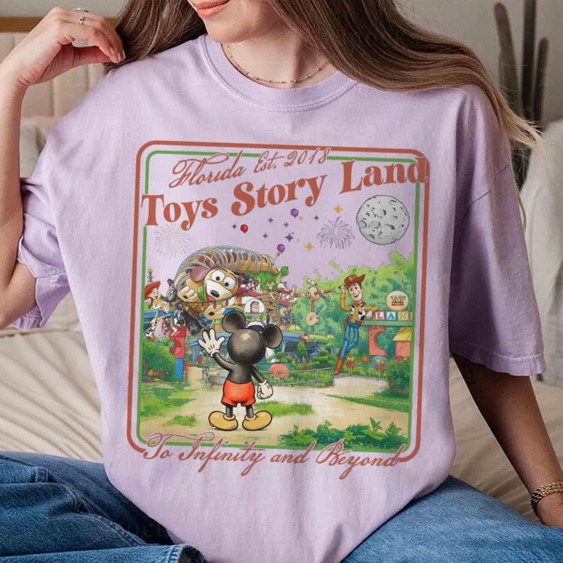 May include: A lilac t-shirt with a vibrant graphic depicting a theme park scene with Mickey Mouse, Woody, and other characters. The graphic includes the text "Florida Est. 2018 Toys Story Land" and "To Infinity and Beyond."