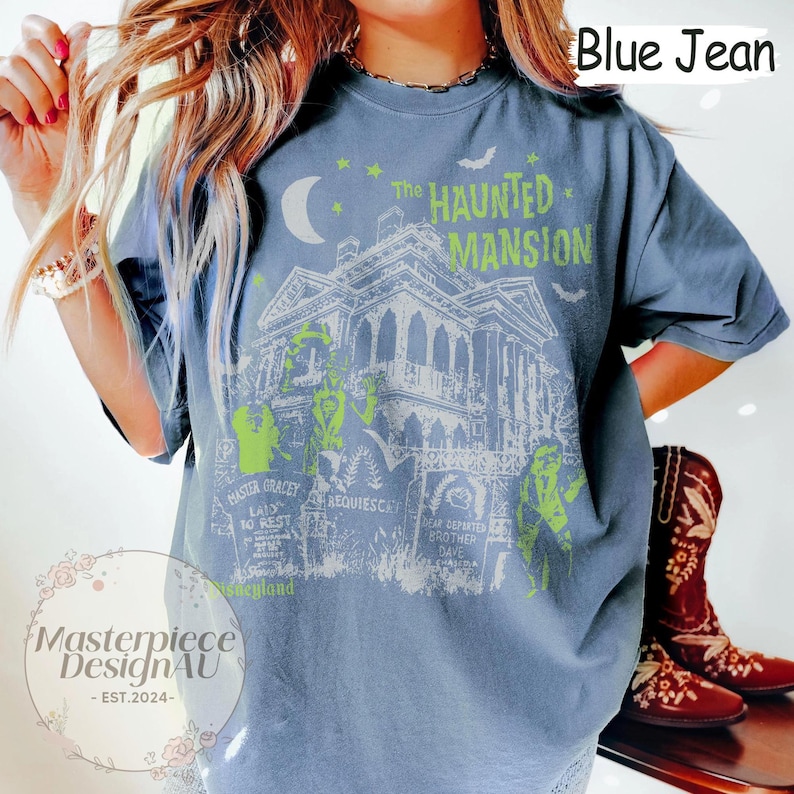 May include: Blue jean-colored t-shirt featuring a graphic of The Haunted Mansion. The design includes the text "The Haunted Mansion" and various figures in neon green. The shirt also has the text "Masterpiece DesignAU - EST.2024-".