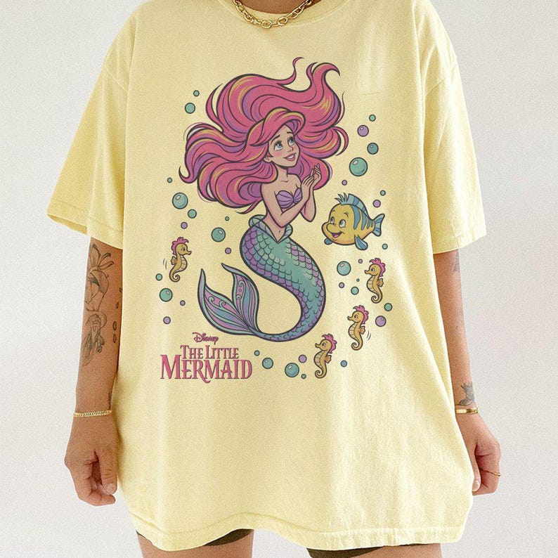 May include: A pale yellow t-shirt featuring a graphic print of Ariel from The Little Mermaid with long pink hair and a teal tail. The design includes Flounder, several seahorses, and bubbles, with the text Disney The Little Mermaid printed below.