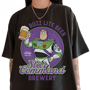 May include: Black t-shirt featuring a graphic of Buzz Lightyear holding a beer mug, with the text "Buzz Lite Beer" and "Star Command Brewery." The design includes a purple circle with green and white accents, and the words "Best Quality Since 1995."