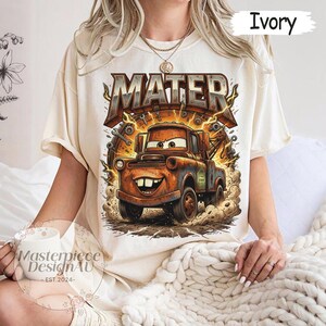 May include: Ivory-colored t-shirt featuring a cartoon image of Mater from the movie Cars. The graphic includes the name "MATER" in a distressed, brown font with lightning bolts. The shirt is a crew neck and has short sleeves.