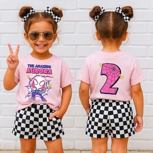 May include: A young child wearing a pink t-shirt with the text "THE AMAZING AURORA" and a cartoon graphic. The child is also wearing black and white checkered shorts and has two buns with checkered bows.