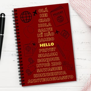 May include: A red spiral-bound notebook with the word "HELLO" in yellow and travel-themed illustrations. The notebook features greetings in various languages, including "OLÁ," "CIAO," and "BONJOUR."