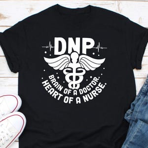 Può includere: T-shirt nera con grafica bianca con le lettere "DNP" sopra un simbolo di caduceo alato. Il testo "Brain of a Doctor, Heart of a Nurse" è sotto. Scarpe da ginnastica bianche visibili nell'angolo in basso a sinistra.