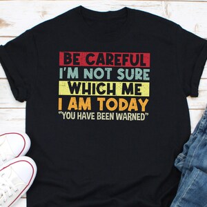 May include: Black t-shirt with the text "BE CAREFUL I'M NOT SURE WHICH ME I AM TODAY YOU HAVE BEEN WARNED" in red, yellow, and blue block letters. The shirt is paired with white sneakers and blue jeans.