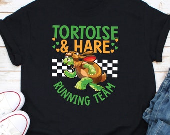 Op de afbeelding: Zwart T-shirt met een cartoonafbeelding van een schildpad verkleed als een konijn, met de tekst "TORTOISE & HARE RUNNING TEAM". Het ontwerp bevat groene en oranje harten en een geblokte vlag.