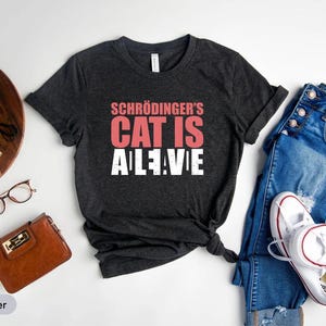 Könnte beinhalten: Dunkelgraues T-Shirt mit dem Text "Schrödinger's Cat Is Alive" in Rosa und Weiß. Das Shirt wird mit blauen Jeans und weißen Turnschuhen kombiniert, zusammen mit Accessoires wie Brille und Geldbörse.
