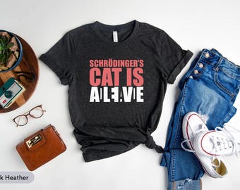May include: Dark heather gray t-shirt with the text "Schrödinger's Cat Is Alive" in pink and white. The shirt is paired with blue jeans and white sneakers, with accessories like glasses and a wallet.