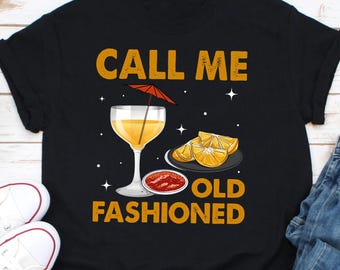 Puede incluir: Camiseta negra con el texto "CALL ME OLD FASHIONED" en dorado. El gráfico incluye un cóctel con sombrilla, rodajas de naranja y un plato pequeño con salsa roja. Zapatillas blancas y vaqueros azules son visibles.
