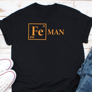 May include: Black t-shirt with the text "Fe MAN" in orange. The "Fe" is in a box with the atomic number 26 and the atomic mass 55.85. White sneakers and blue jeans are visible in the background.