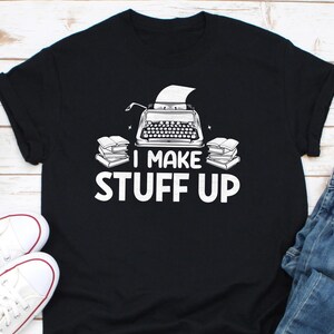以下が含まれることがあります： 白いタイプライターと積み重ねられた本のグラフィックが描かれた黒いTシャツ。グラフィックの下に「I make stuff up」というテキストが印刷されています。