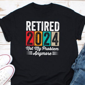 Puede incluir: Camiseta negra con el texto "RETIRED 2024 Not My Problem Anymore" en blanco, rojo, naranja, turquesa y beige. El diseño es de estilo retro con un aspecto desgastado. La camiseta se combina con zapatillas blancas y vaqueros azules.