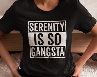 May include: Black t-shirt with the text "SERENITY IS SO GANGSTA" in a white, distressed font within a white rectangular frame. The shirt is short-sleeved and appears to be made of a soft material. The person is wearing black pants.