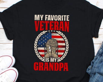 May include: Black t-shirt featuring the text "MY FAVORITE VETERAN IS MY GRANDPA" in white and red. The design includes an American flag, a military helmet, and boots inside a wreath.