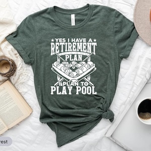 May include: Heather forest green t-shirt with the text "YES I HAVE A RETIREMENT PLAN I PLAN TO PLAY POOL" in white. The graphic features a pool table with billiard balls and crossed pool cues.