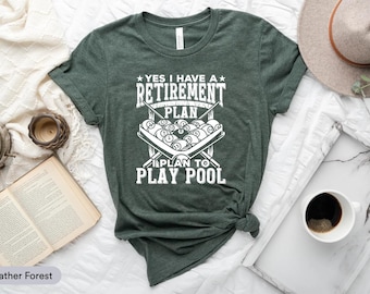 May include: A heather forest green t-shirt with the text "YES I HAVE A RETIREMENT PLAN I PLAN TO PLAY POOL" in white. The graphic features a pool table with billiard balls and crossed pool cues.