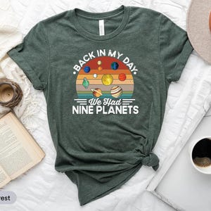 May include: A heather forest green t-shirt with a retro graphic of planets and the text "Back in my day. We had nine planets." The design features planets in various colours against a sunset background.