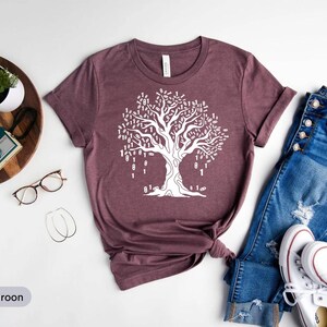 A casual outfit consisting of a maroon t-shirt with a white tree design, blue jeans, and white sneakers, along with a pair of glasses and a potted plant, is laid out on a white background.